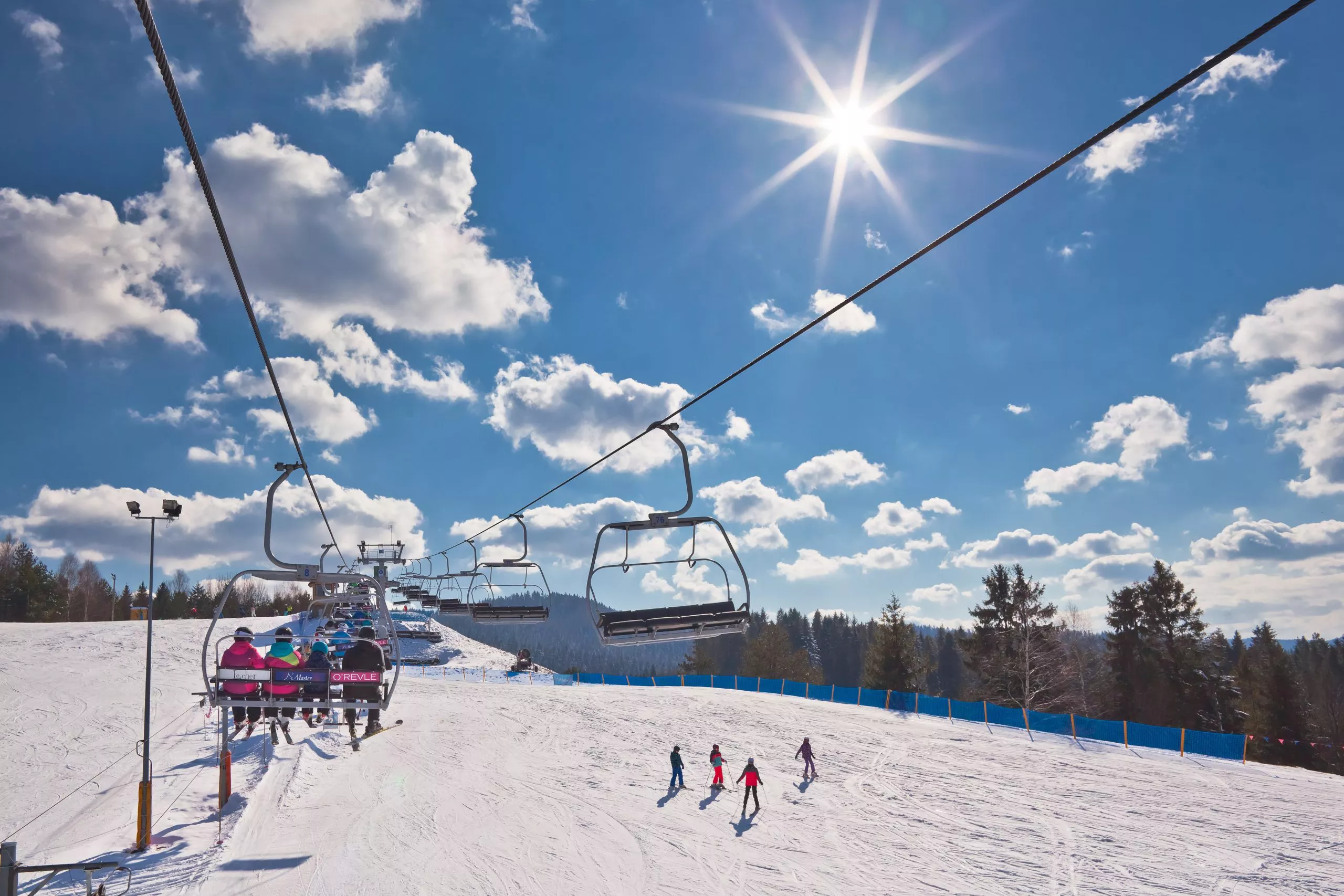 Four small silhouettes on a snowy piste centre bottom; on the left, a chair ski lift takes other skiers to the top; most of the picture is taken by the skies with some clouds and the sun.