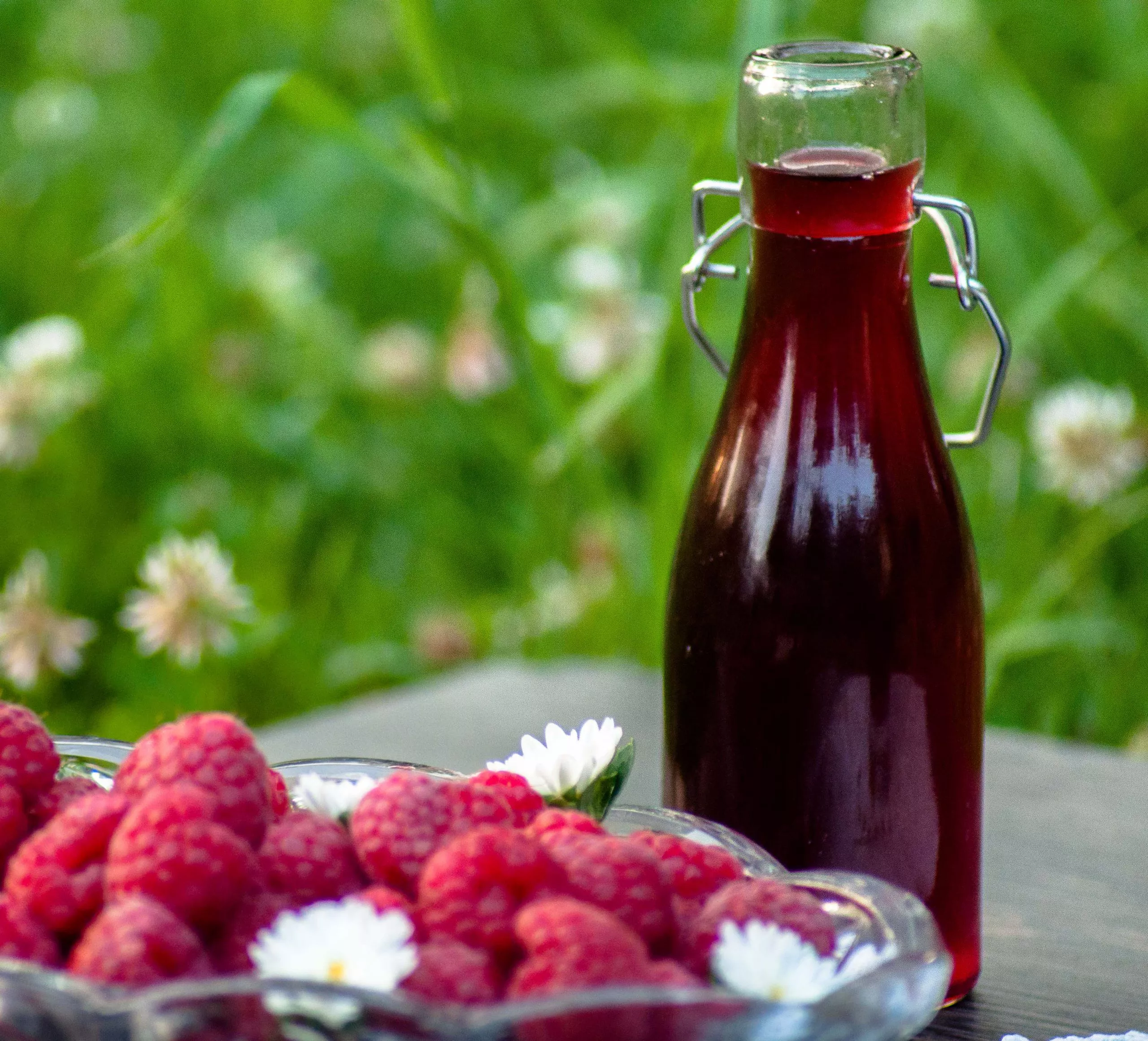 Bottle full of raspberry syrup behind a plateful of ripe raspberry decorated with three daisies; blurred green grass in the background.