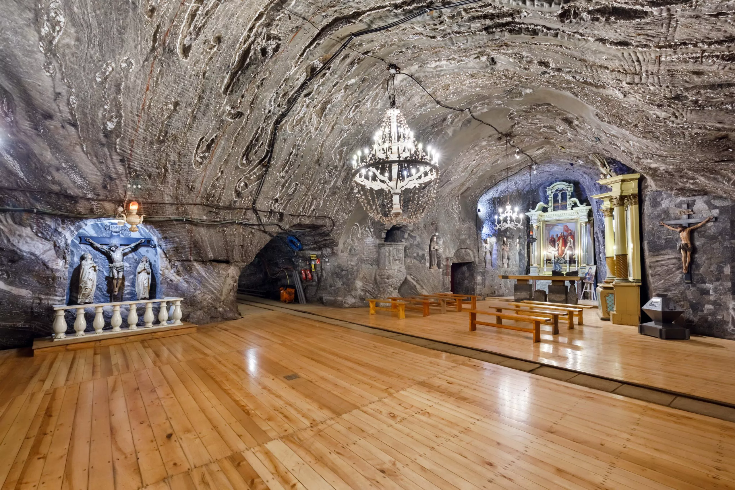 Chapel of St Kinga in a salt mine: an expanse of polished wooden floor under a swathe of stripped grey rock salt forming the walls and ceiling, from which an impressive chandelier hangs. The chapel is furnished with a small crucifixion scene on the left and a prayer area with wooden benches in front of an altar arranged in a niche on the right.
