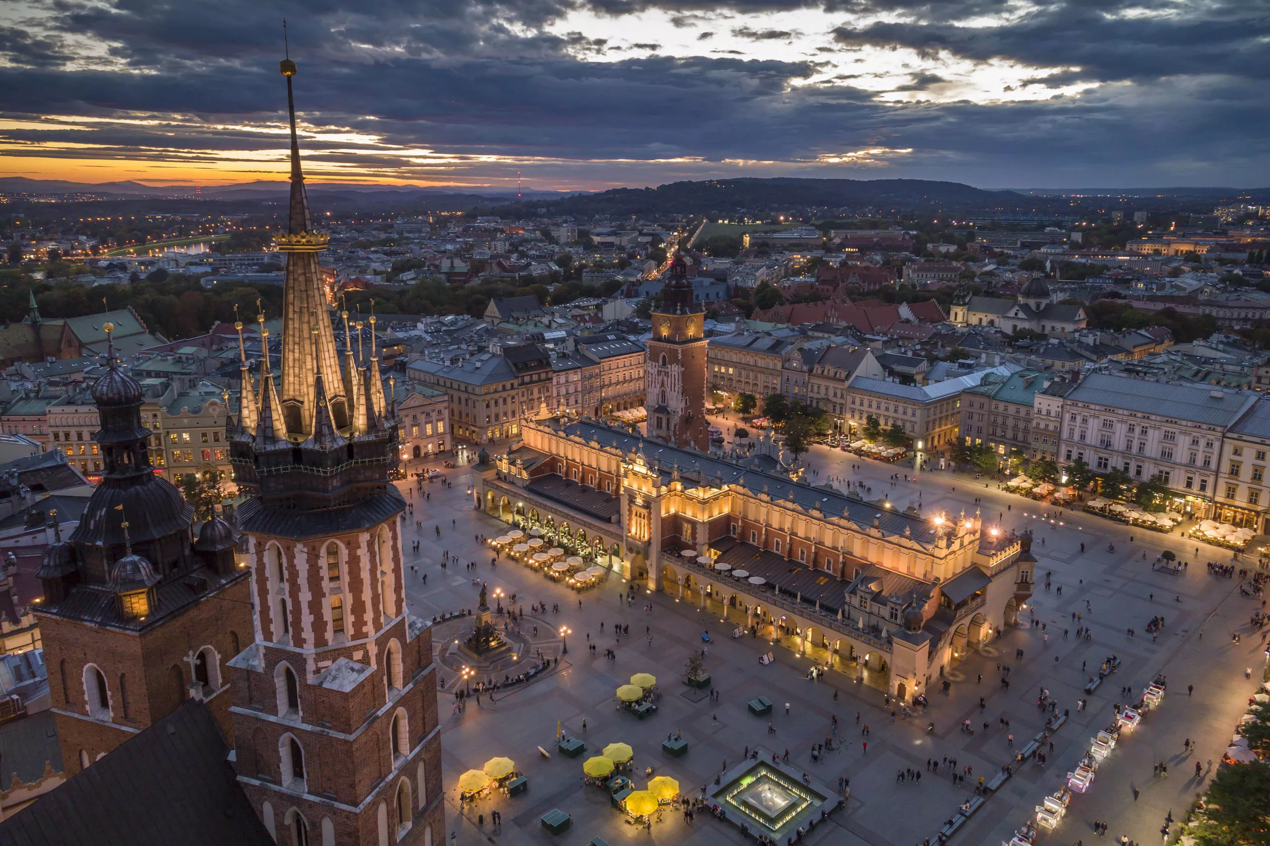 A night-time bird’s eye view of the historical city centre of Kraków. The two towers of unequal height of St Mary’s visible in the foreground on the left stand against a broad background of artificially lit Main Market Square. Standing around it are mansions and townhouses, and its centre is taken by the long Cloth Hall and the Town Hall Tower. Above, the sun is setting below dark clouds.