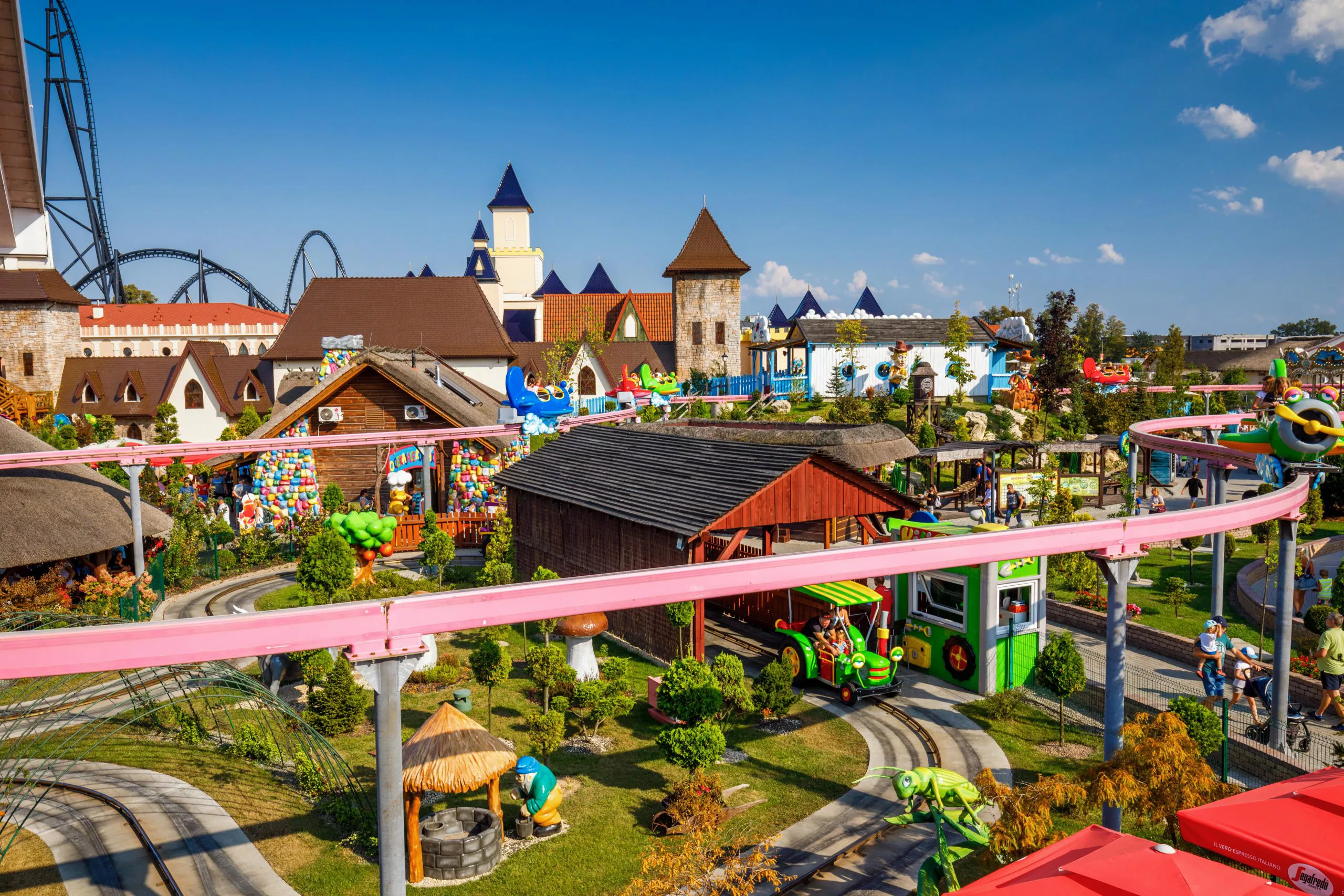 Colourful attractions under a pink monorail in an amusement park include green tractors on a track among other colourful attractions, with a mockup of a medical city and rollercoasters tracks behind.