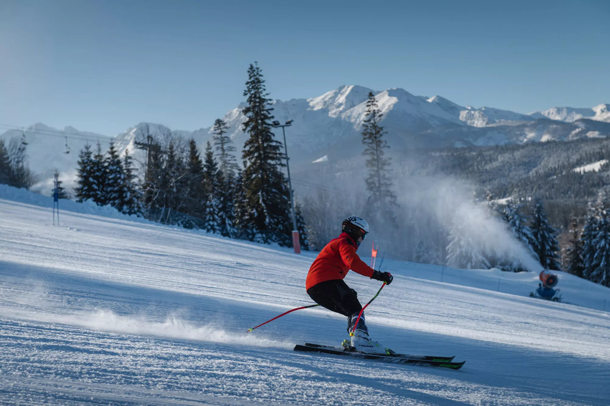 Blick auf einen Skifahrer, der die Piste hinunterfährt. In der Ferne sieht man eine Schneekanone, einen Sessellift, Bäume und die verschneite Tatra. Der Himmel ist wunderschön blau und wolkenlos.