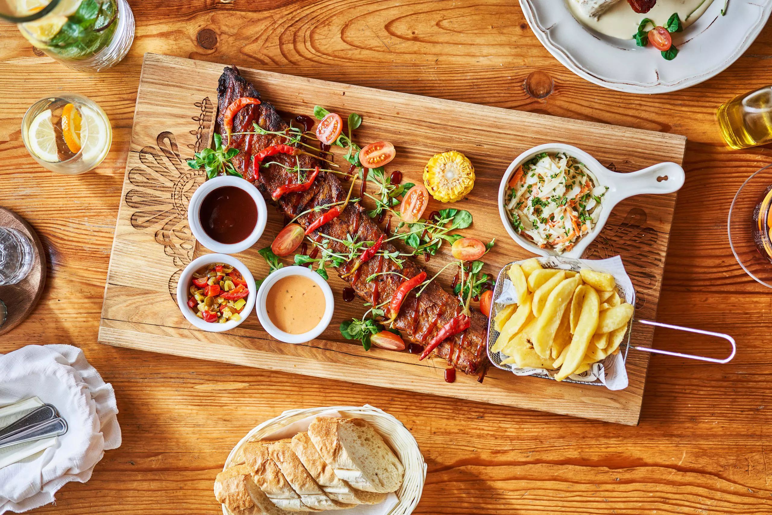 Top view of a wooden board with garnished spareribs, with sides and sauces; other dishes arranged around.