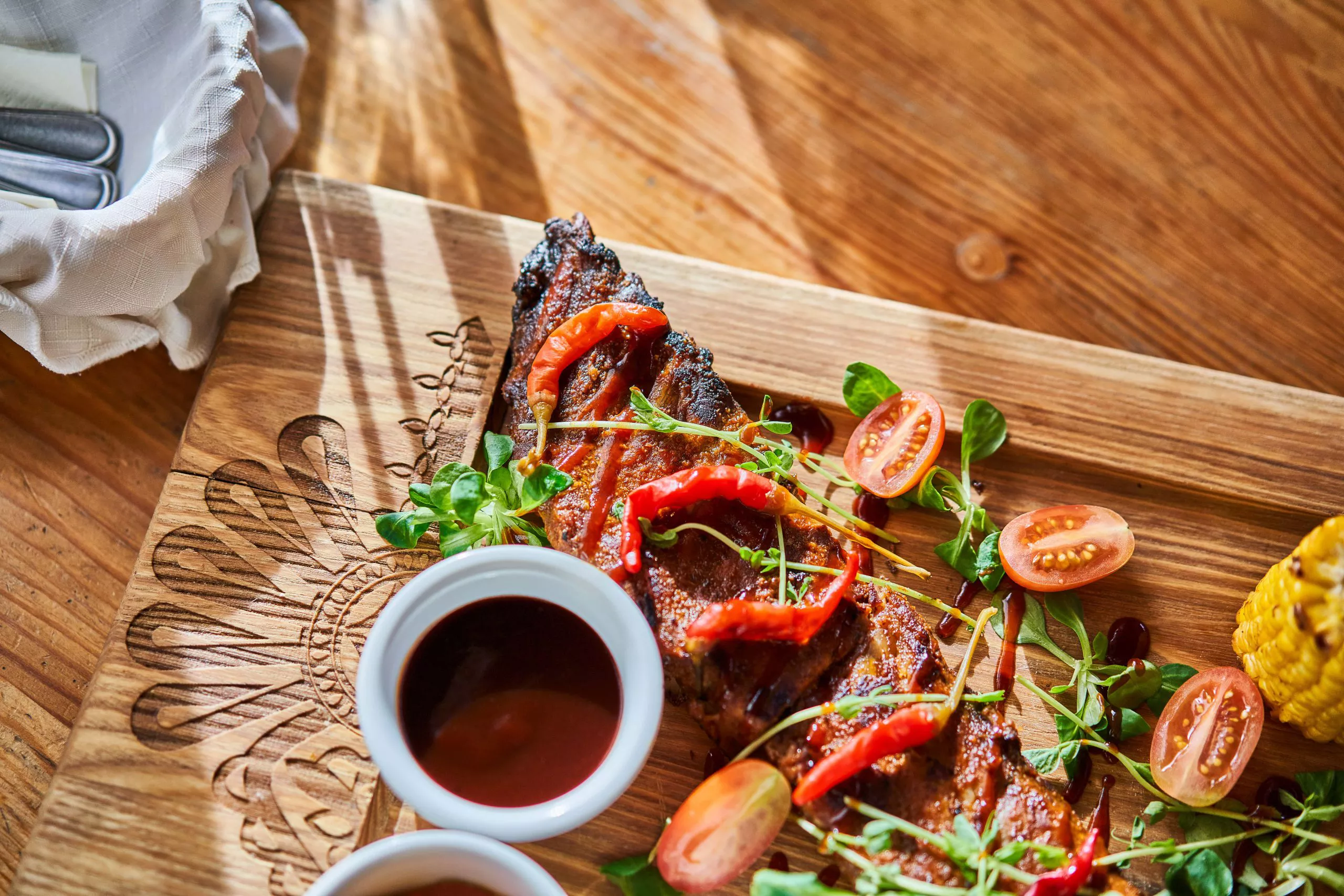Pork ribs garnished with chilis, cherry tomatoes, and lamb’s lettuce leaves, are arranged on a wooden board, its Polish highland ornamentation partly covered by ramekins with sauce. A corner of a cloth-lined basket with some cutlery was caught in the top left-hand corner of the shot.
