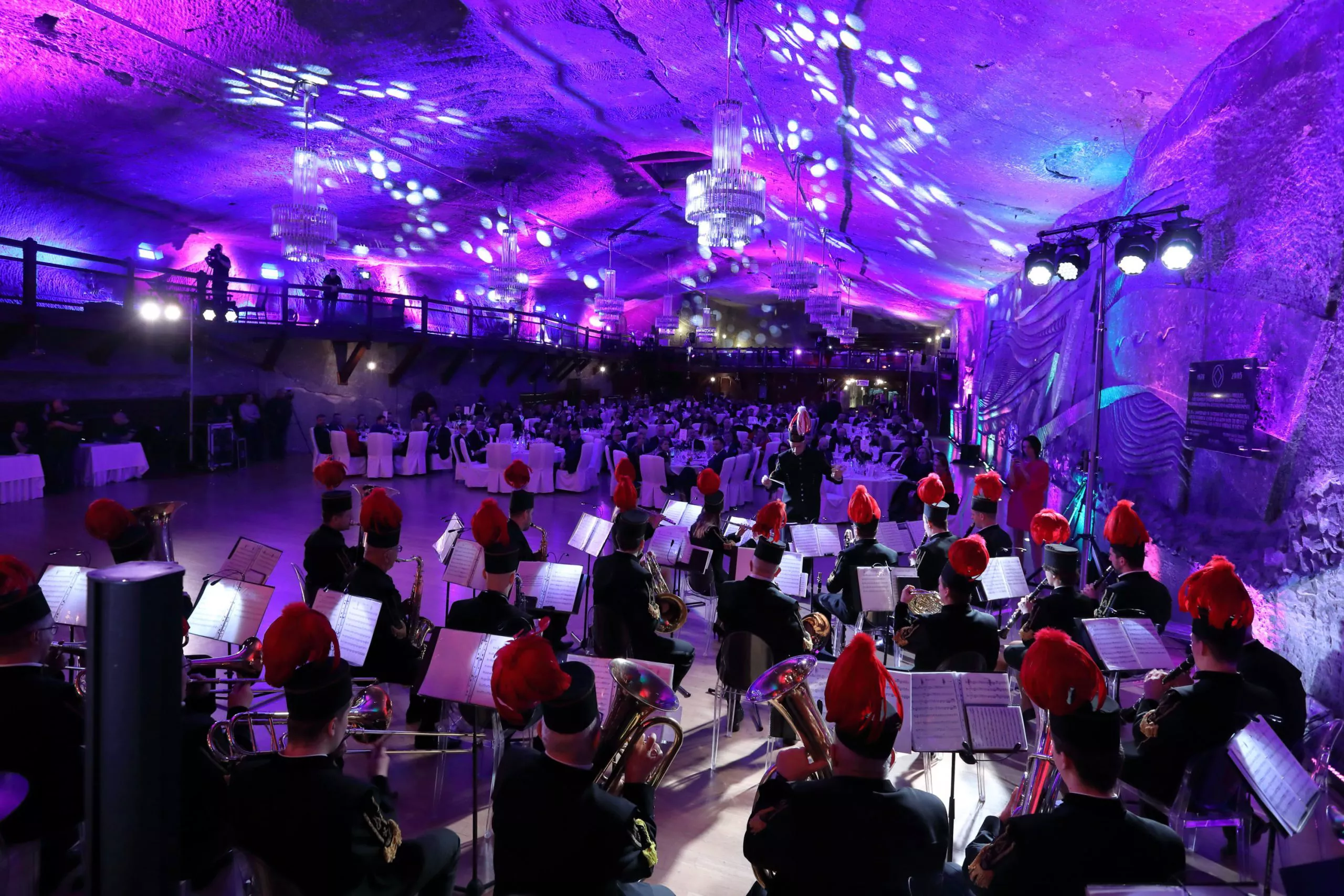 An event in a grand subterranean hall seen from behind the orchestra in traditional mining dresses including tall black caps with red plumes.