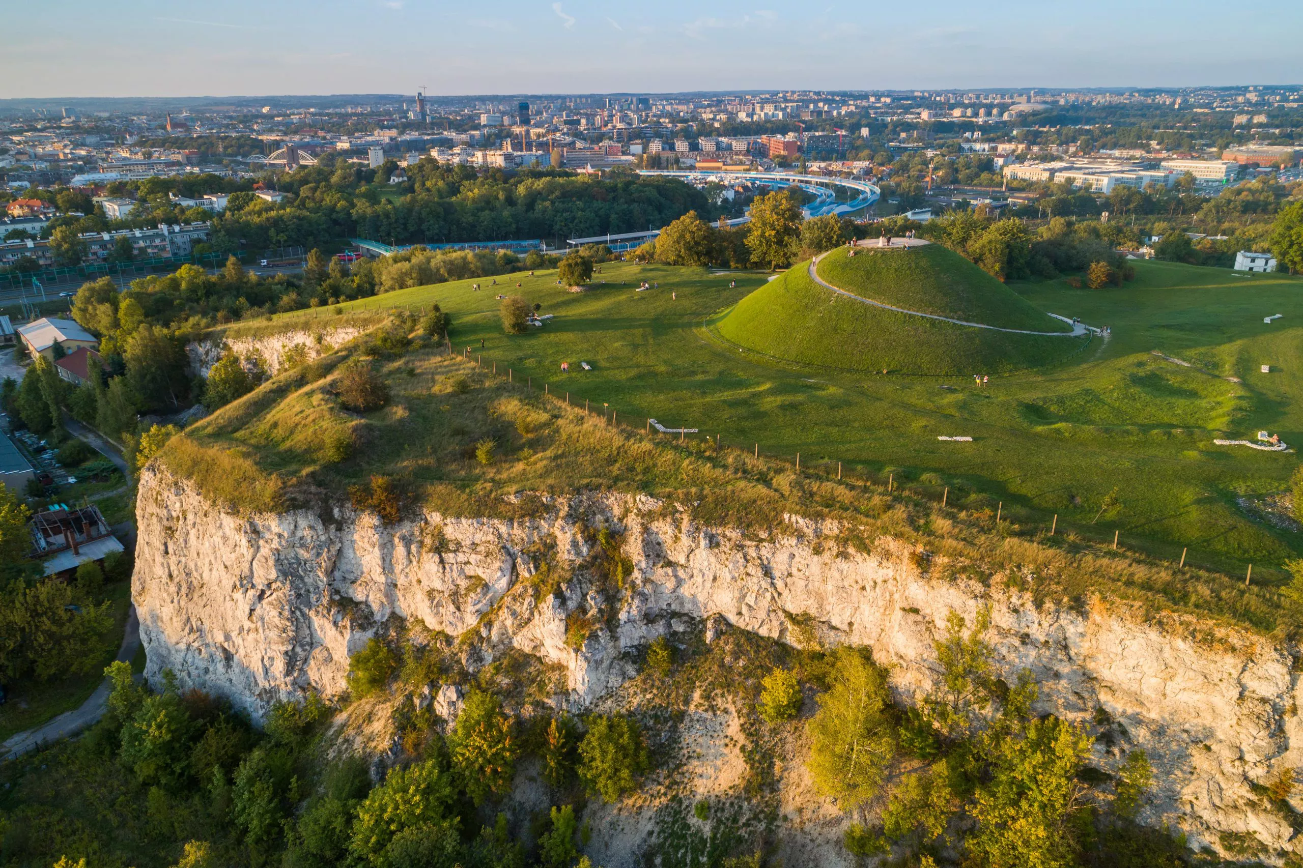 In this aerial view, Mound of King Krak (Krakus) stands among the green turf on the left, above the vertical limestone cliffs of Liban Quarry. The mottle in the distance are the eastern districts of Kraków, visible under an overcast sky.