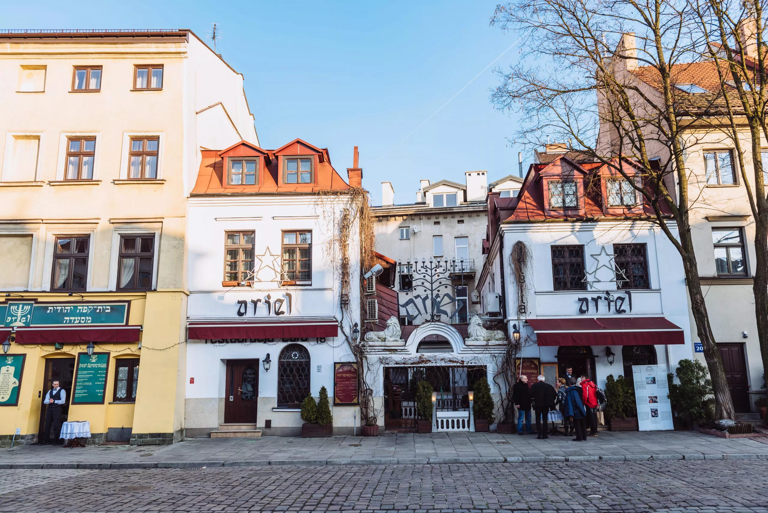 Two almost identical small three-storied white buildings with red roofs flank a low gate of masonry topped by two white lions, huddling between two taller buildings. The name “Ariel” displayed on all the three central structures is that of a popular Kazimierz restaurant. A group of tourists standing outside on the pavement under a tall leafless tree are greeted in its door by a waiter.