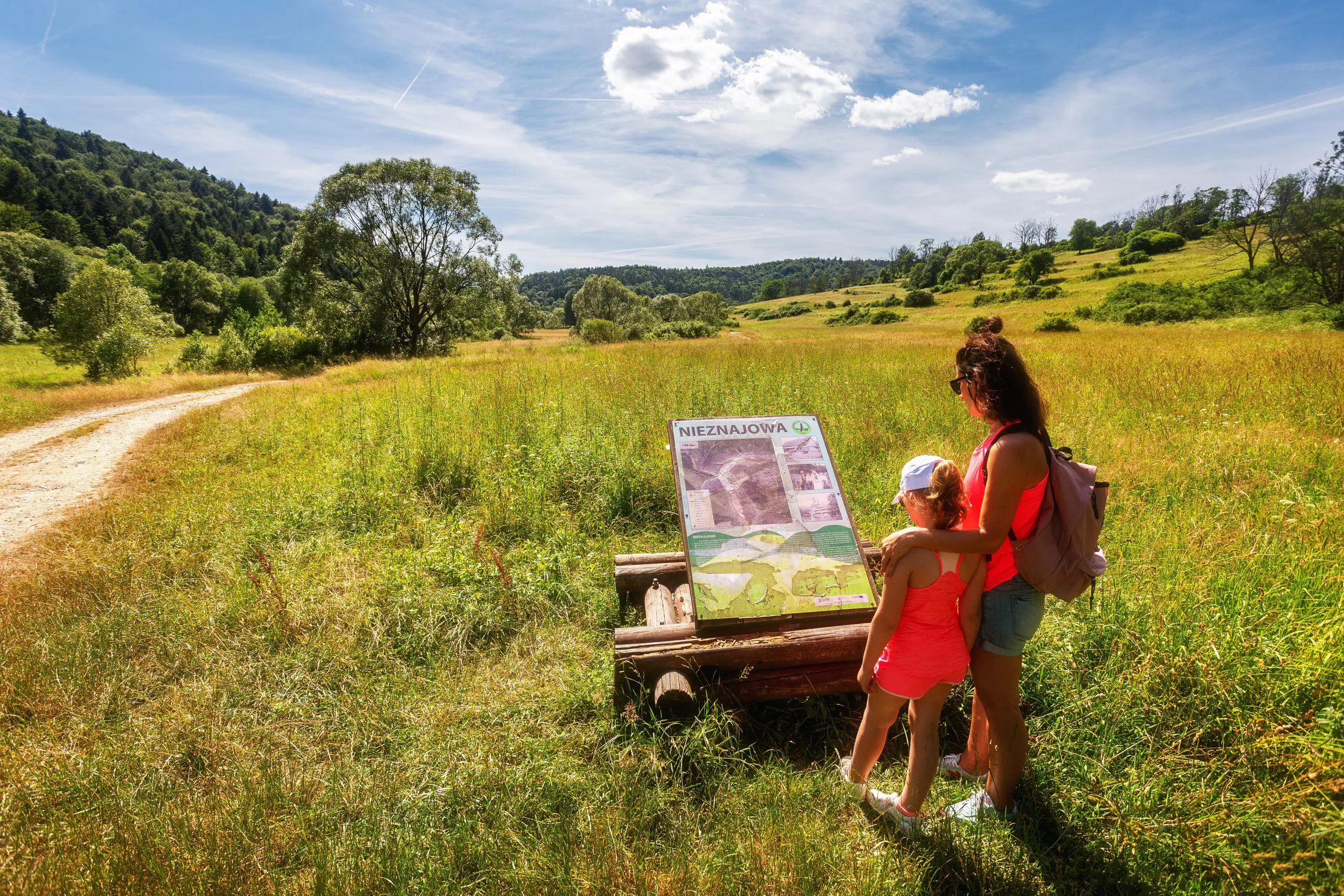 Young woman and little girl on a summer trail read information on a board mounted in a meadow, the dirt road on the left runs towards clusters of trees and hills on the horizon under cloudy blue skies.