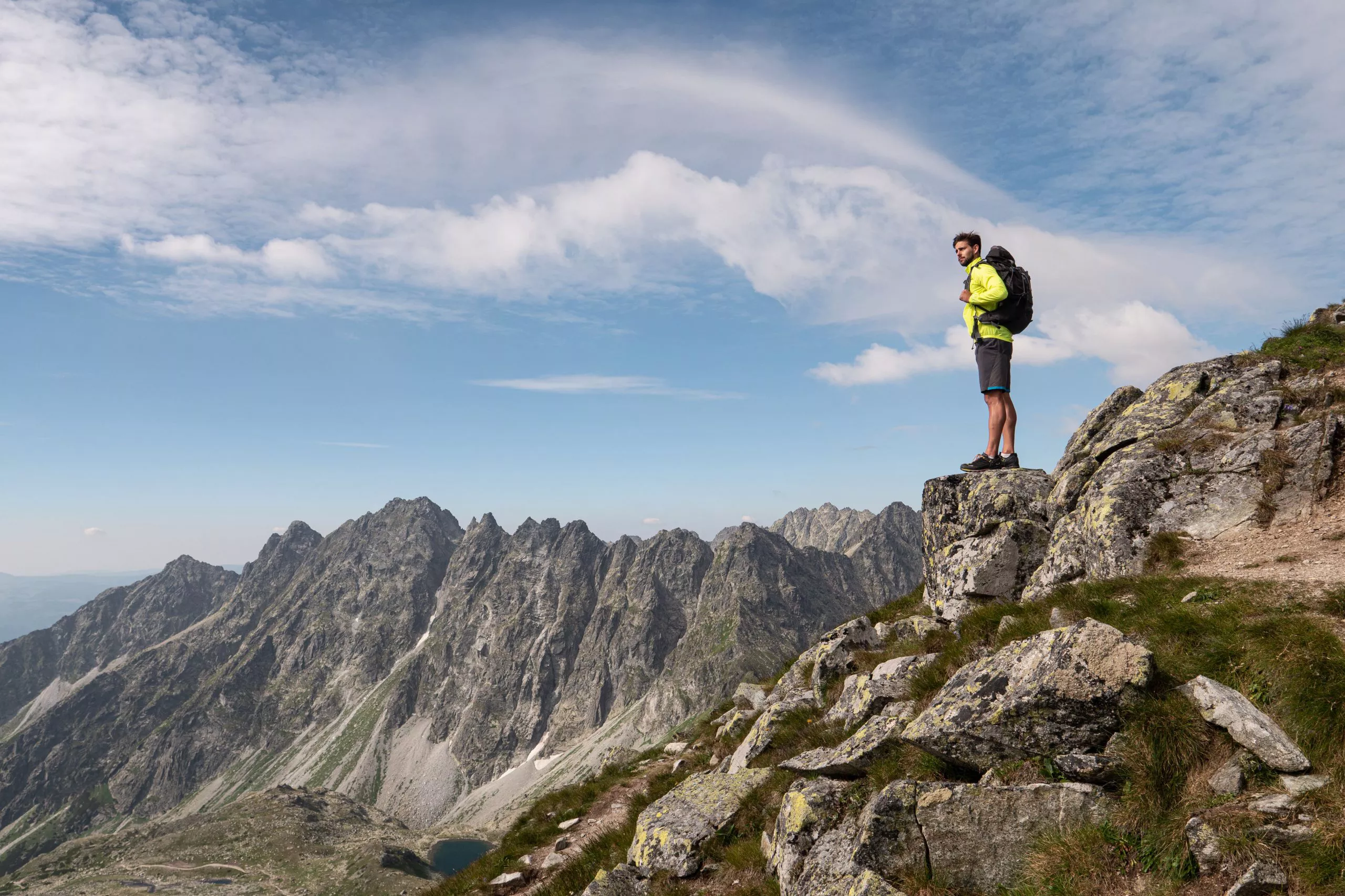 Blick auf einen jungen Mann mit einem großen Rucksack. Der Mann steht auf einem Felsen und schaut in die Ferne. Ringsherum sind die Gipfel der Tatra zu sehen, im Tal ist das Fragment eines Sees zu erkennen, und in den Rinnen sind Spuren von Schnee zu sehen. Der Himmel ist wunderschön blau, von mittelgroßen weißen Wolken bedeckt.