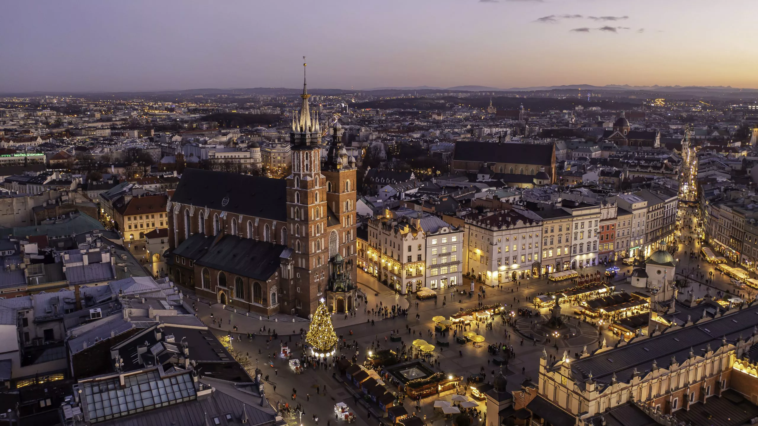 The eastern half of the artificially lit Main Market Square proudly demonstrates its Christmas lighting at pre-dusk. The bottom left-hand corner is taken by the roof of the Cloth Hall. A tall Christmas tree is standing to its left in front of the uneven-towered St Mary’s Church. A large expanse of the city spreads behind a line of bright façades of Main Market Square mansion to its right, reaching the horizon behind which the Sun has just set.