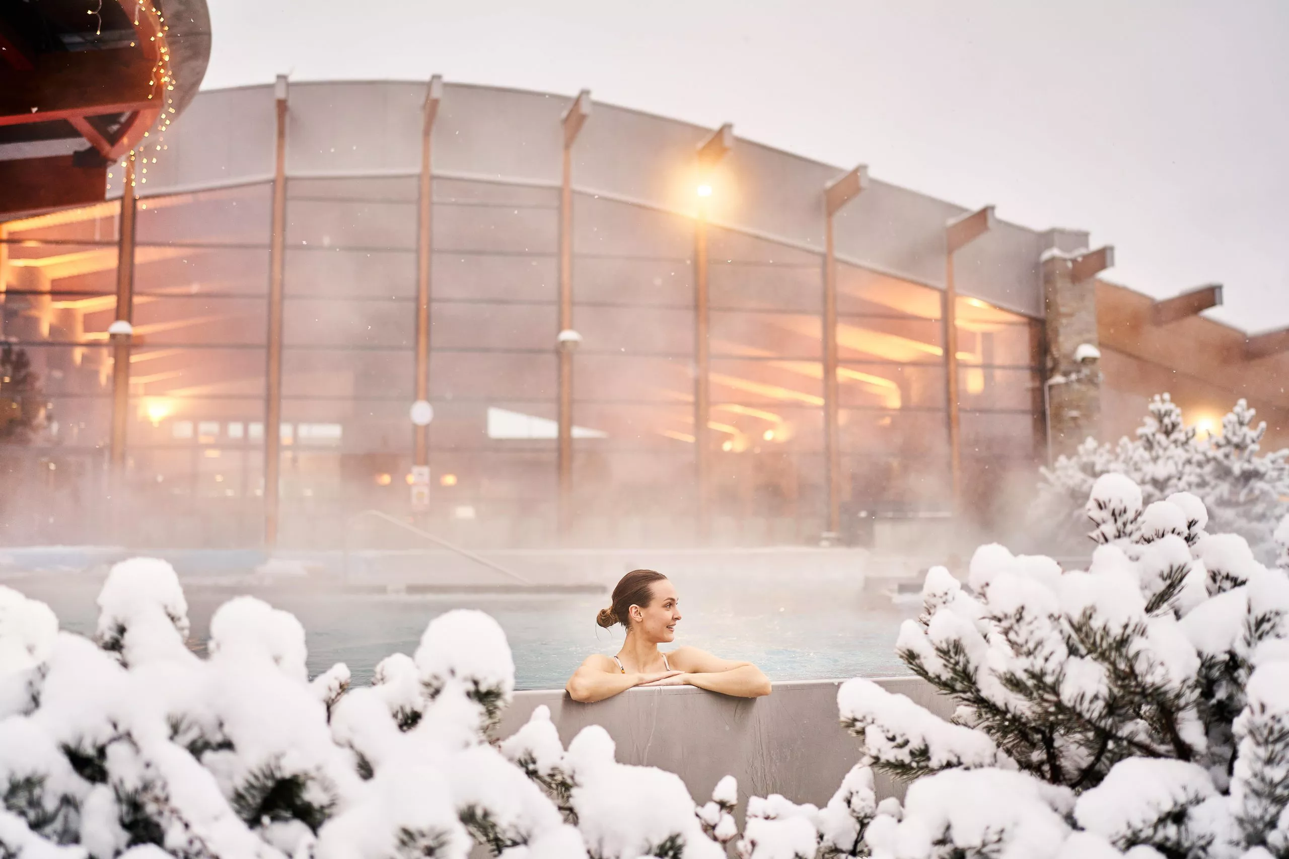 Blick auf eine junge Frau im Thermalbad, mitten verschneiter Bäume. Im Hintergrund ist das beleuchtete Gebäude des Thermalbads zu sehen. Aus dem Becken steigt heißer Dampf auf.