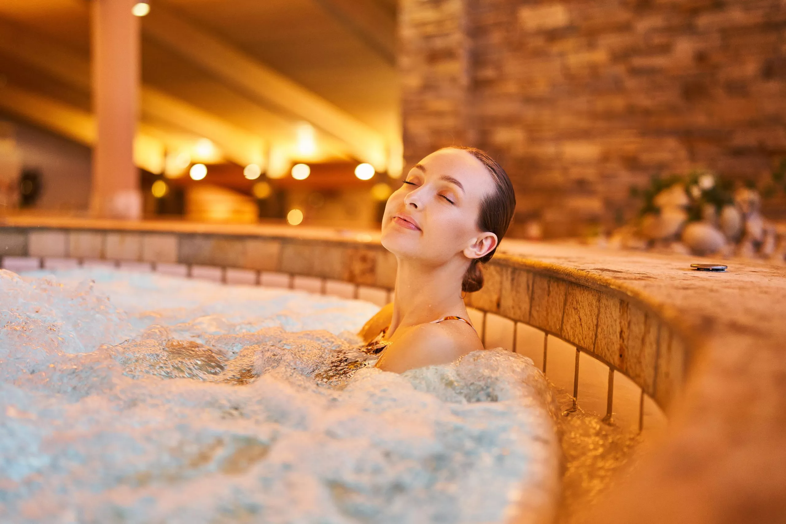 Blick auf eine junge, entspannte Frau mit geschlossenen Augen, die in einem Becken mit sprudelndem Wasser sitzt. In der Ferne sieht man die Beleuchtung und das steinige Innere des Pools: Wände, Decken.