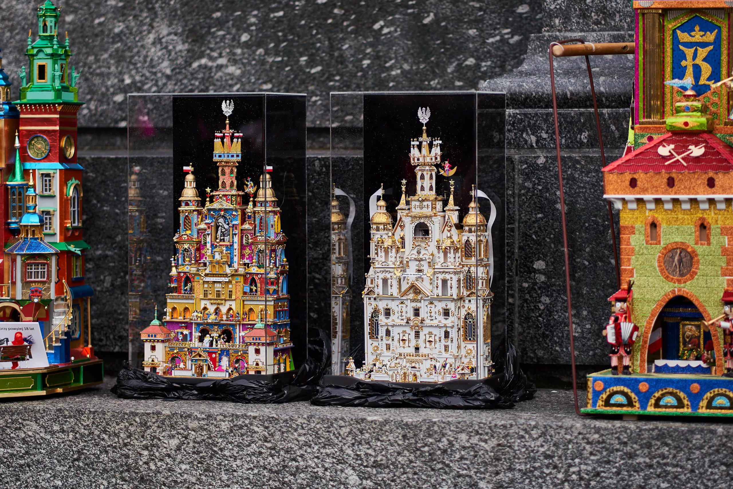 Four miniature architectonic constructions composed of various elements of Kraków architecture with centrally placed miniature scenes presenting nativity of Jesus, all rich in detail and covered in extremely colourful tinfoil, stand on a dark stone ledge against the background of mottled black marble.