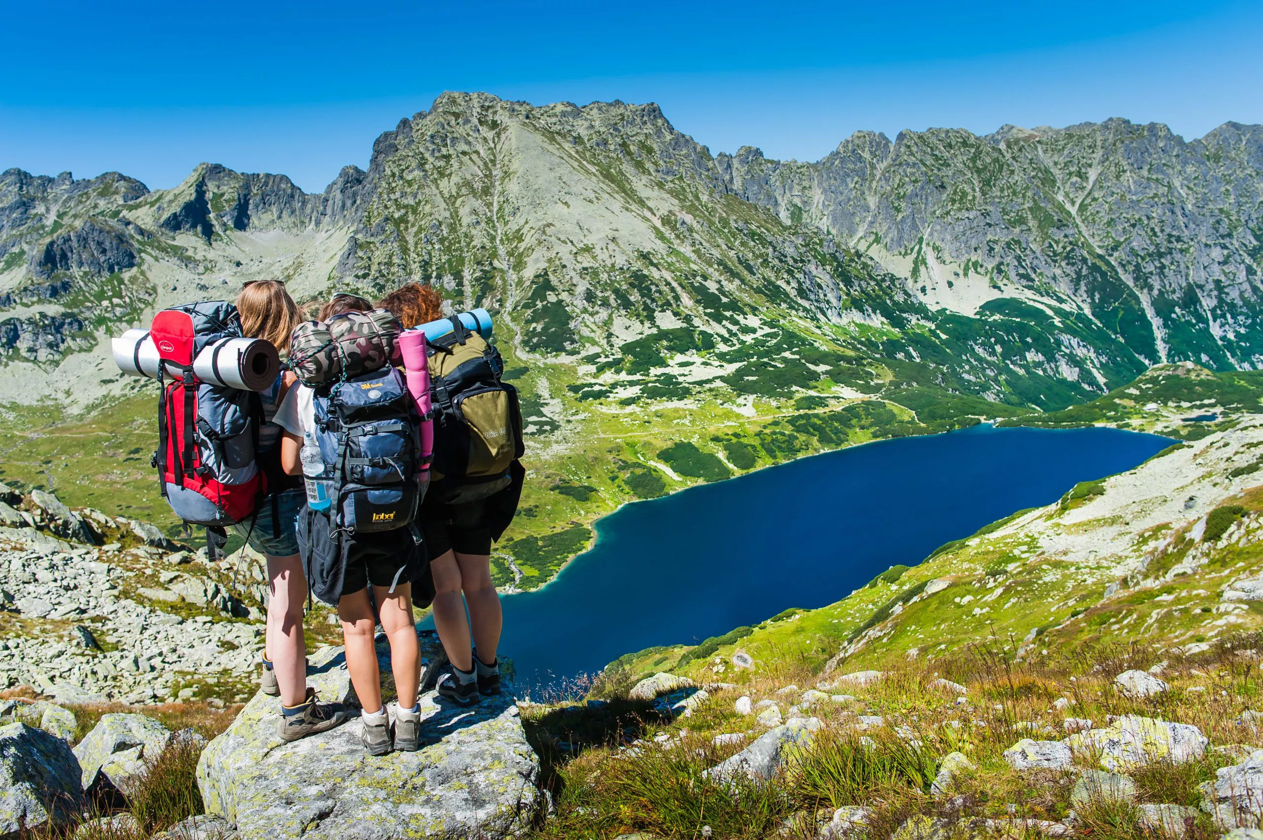 Three girls carrying large backpacks stand on a rock on the left and look down into a valley with a dark blue mountain lake (tarn); its greenish valley is surrounded by a curving range of rocky mountains.