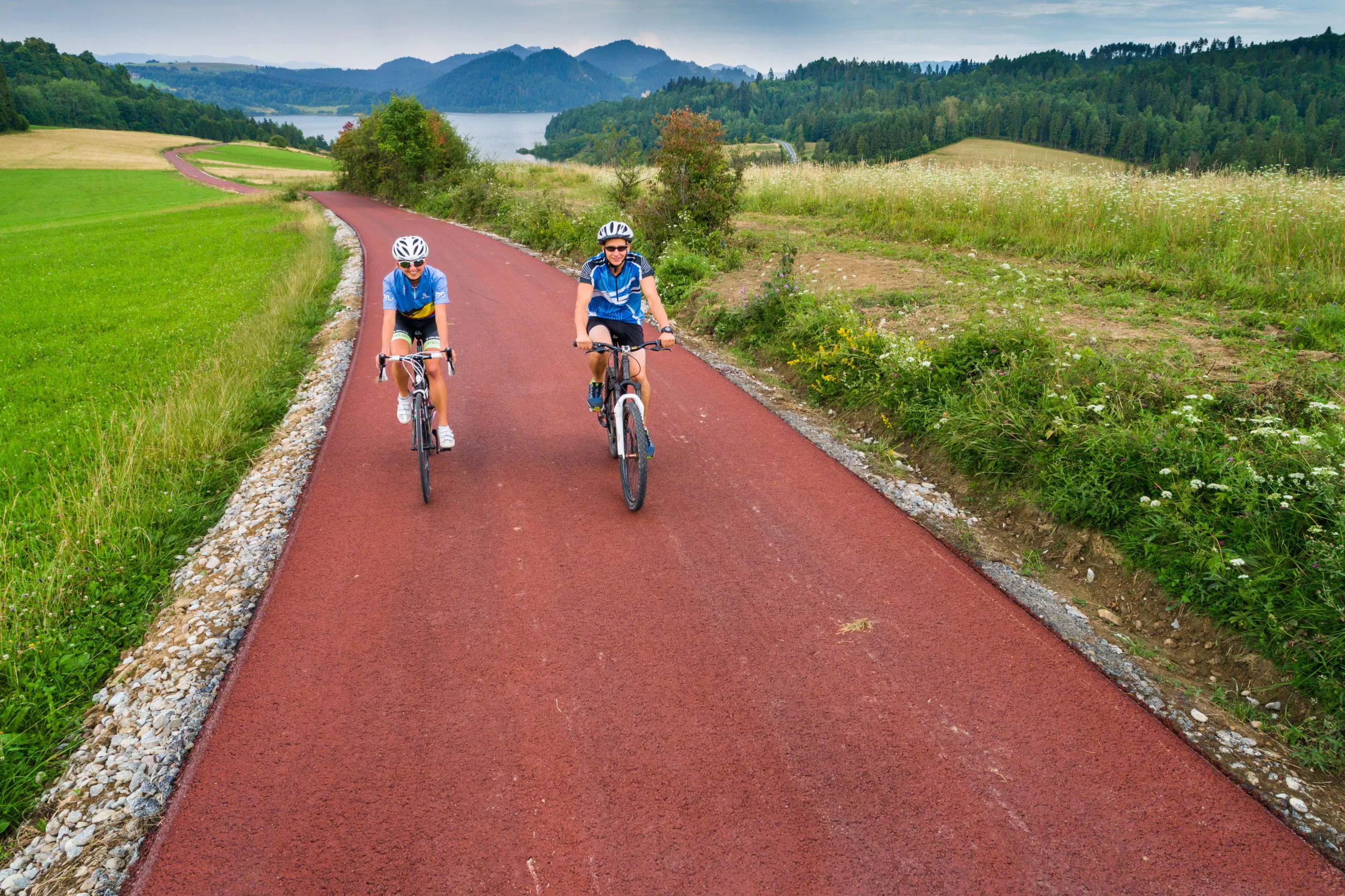 Two people in helmets and blue jerseys ride bikes towards us on a red tarmac cycle path twisting between fields; pale waters of a reservoir surrounded by bluish mountains in the background.