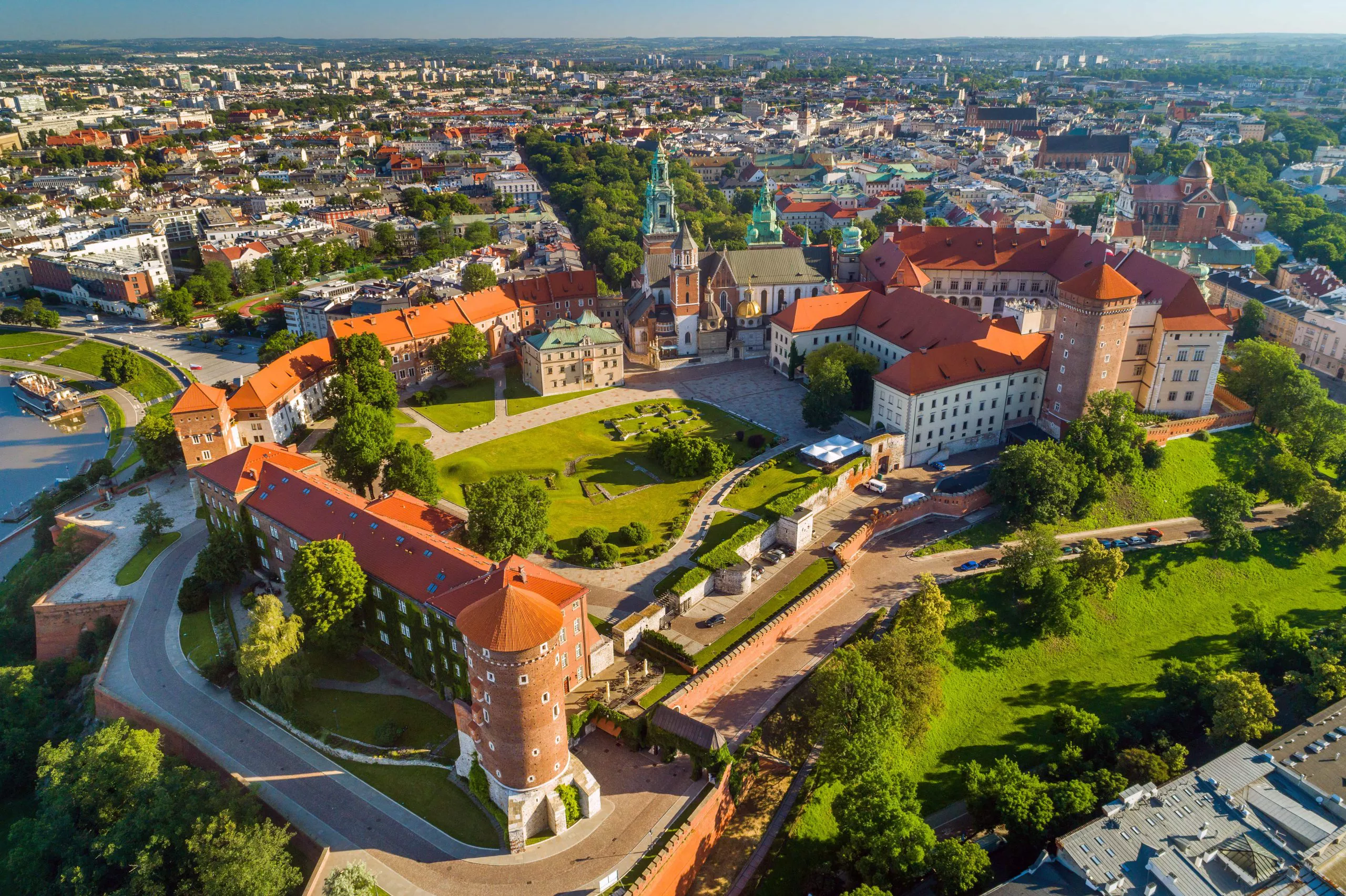 Drohnenansicht des von der Sonne beleuchteten Wawel-Hügels mit der Wawel-Kathedrale, dem Königsschloss, den Türmen und anderen Gebäuden der gesamten Anlage. Einige der Gebäude wurden aus Backstein gebaut. In der Ferne, im Norden, sind Umrisse von Gebäuden und Kirchen in Krakau zu sehen. Auf dem Wawel-Hügel gibt es viele Grünanlagen, darunter Rasen und Bäume.
