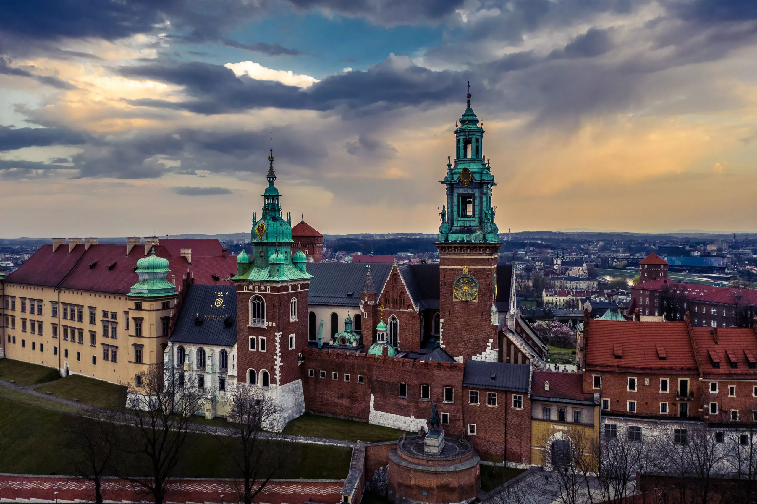 A bird’s eye view of Wawel Hill. The brick and limestone walls of the cathedral with its towers boasting tall patinised spires dwarf both the Gothic houses of the same material on the left, and the wing of the castle plastered white under a tall dark red roof on the left. The distant plane consists of the mottle of buildings under a dusky sky with sandy, blue, white and almost black clouds.