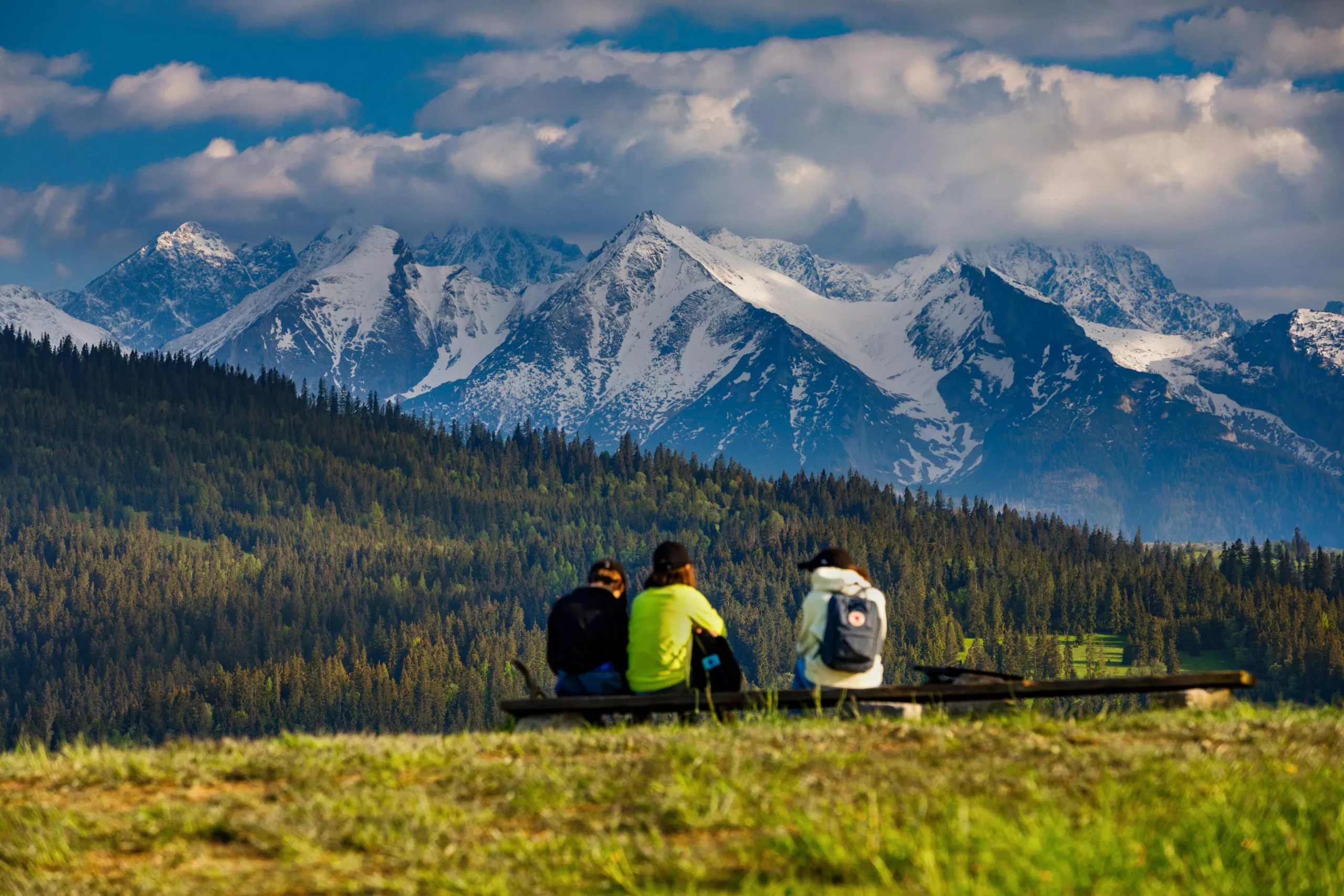 Ein Blick auf drei junge Leute mit Rucksäcken, die auf einer Bank sitzen und die schöne Aussicht auf die schneebedeckten, felsigen Gipfel der Tatra genießen. Unterhalb der Berge befindet sich ein bewaldeter Bereich. Der Himmel ist blau mit vielen weißen Wolken bedeckt, die zum Teil die Berggipfel verdecken.