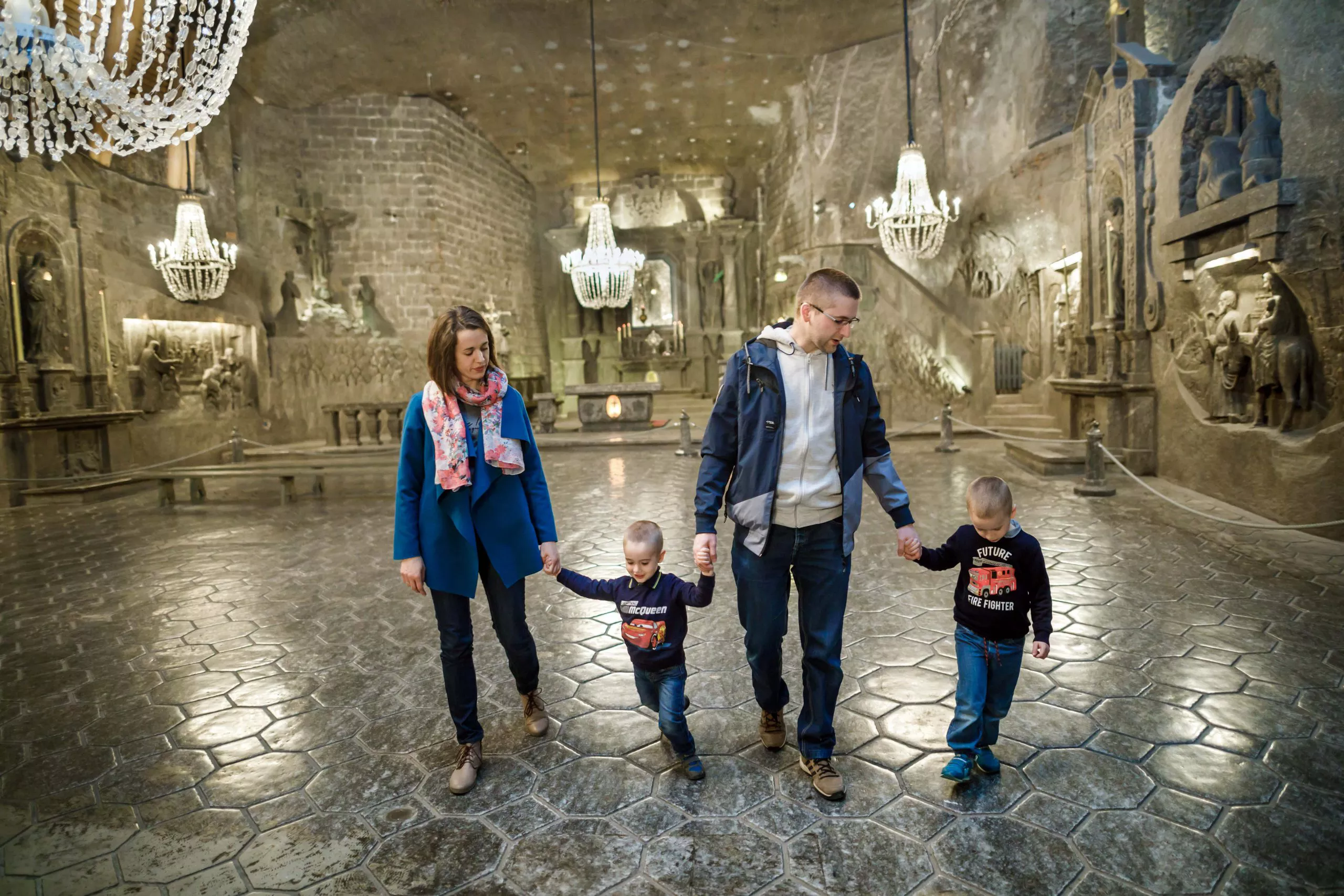 A young family dressed in blue: a woman with a white scarf, a very small boy, a man in a white shirt, and another little boy – all holding hands – walk towards the camera in a spacious underground salt chapel with walls and floor made of rock salt, numerous chandeliers made of salt crystals, and the walls adorned by bass reliefs of religious scenes cut in living salt.