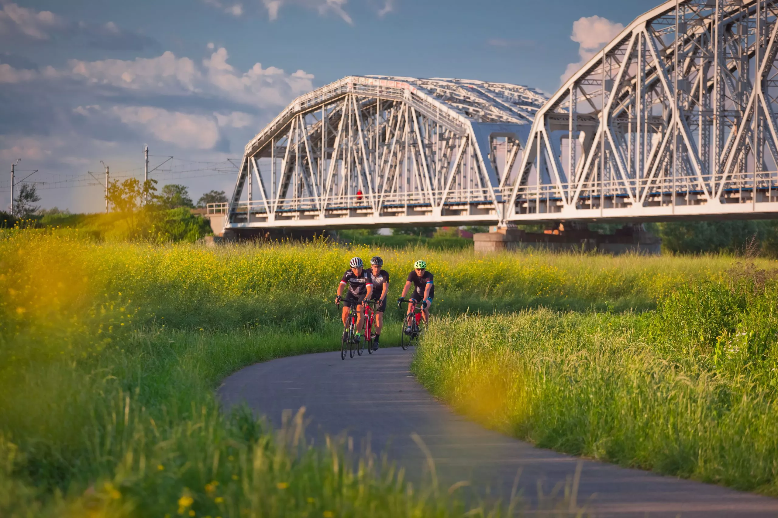 Blick auf einen Abschnitt des Radwegs, auf dem drei junge Radfahrer mit Helmen unterwegs sind. Im Hintergrund ist eine große weiße Brücke über den Weg zu sehen, während ringsherum grünes Gras mit gelben Blumen wächst. Der Himmel ist blau mit mittelgroßen weißen, wogenden Wolken.