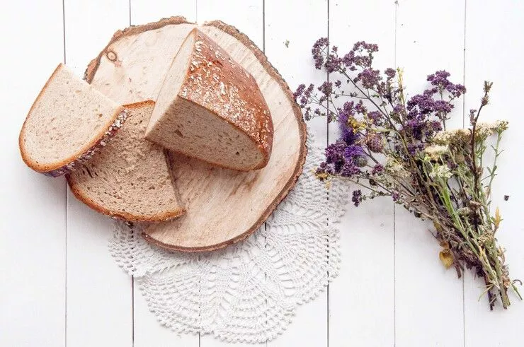 Two slices and a quarter-loaf of Prądnicki bread lie on a large wooden coaster lying on a crochet doily, with a bunch of purple and white flowers of the field to the right.