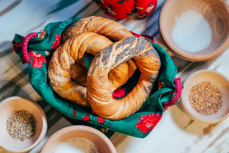 Three rings of parboiled dough baked dark brown are arranged in a basket lined with green cloth decorated with a motif of large red flowers. The basket stands on a wooden table among wooden bowls with salt and sesame seeds which you can also see on the Obwarzanki bread rings.