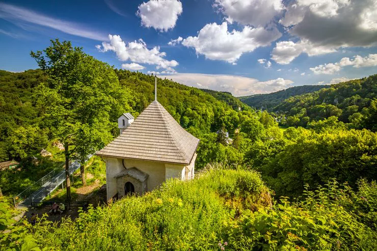 A chapel of bright stone peering from among lush vegetation on a hill ridge. The day is sunny, even though tuft-like clouds in the centre are giving way to darker ones on the right and in the centre, over the distant hills.