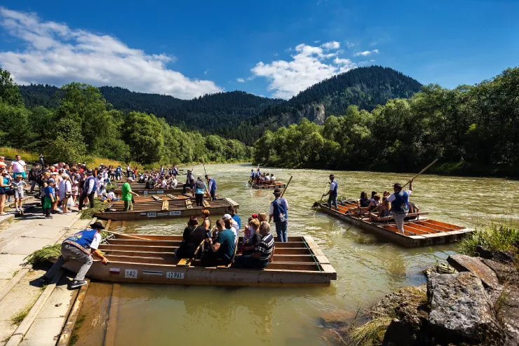 Several dug-out rafts are moored to the shore on the left, waiting for tourists to embark. Two others are already on the river, propelled by raftsmen steering them with long punts. The river runs between two forested banks and further into the wooded rocky hills visible in the background against the slightly cloudy sky.