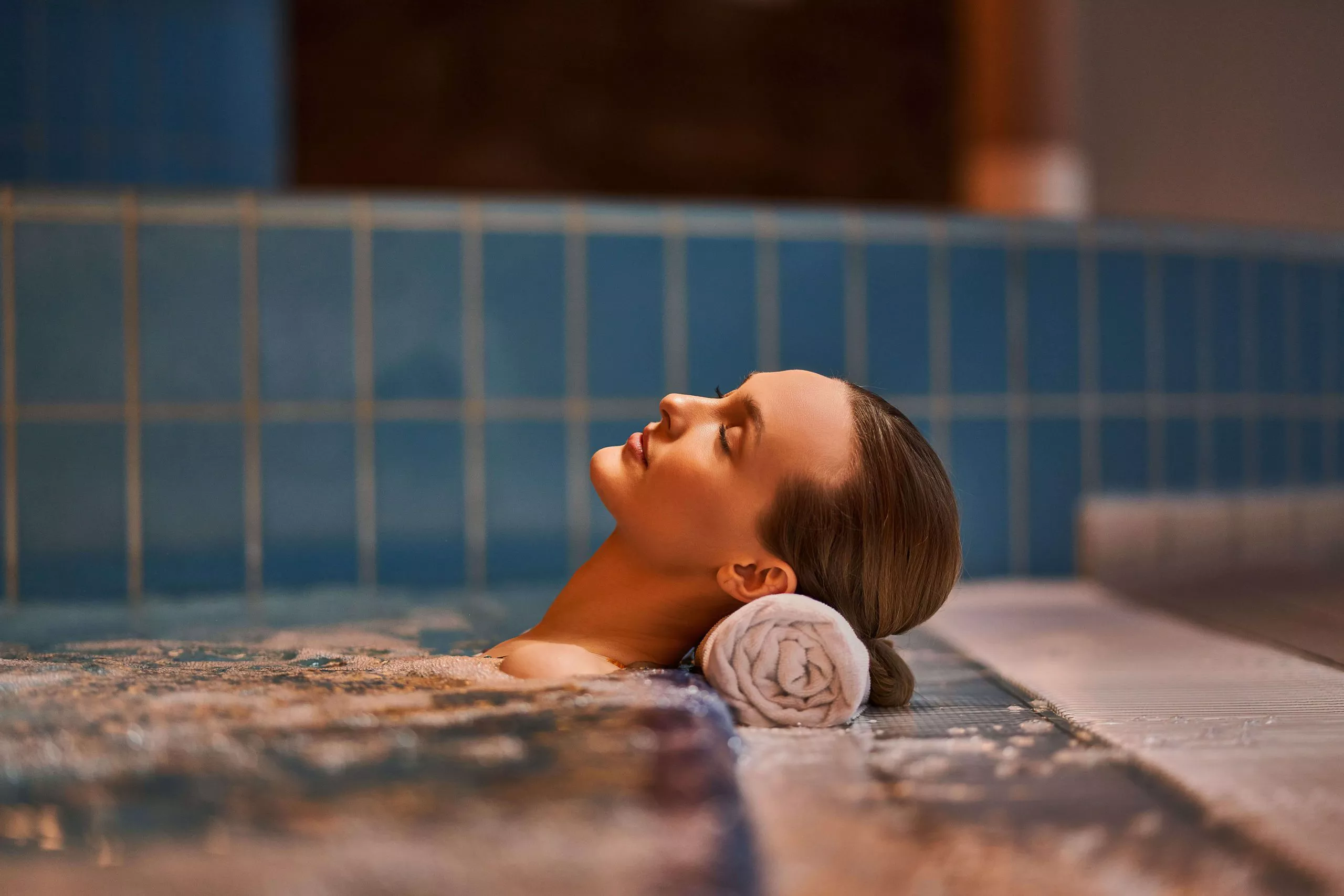A profile view of a woman lying in a pool of water, her eyes closed and her head is gently resting on a rolled white towel. The look of bliss on her face may come from the warmth of the water, in which she is submerged up to the shoulder bone. The blurred blue tiles behind her and a doorway suggest that the photo was taken indoors.