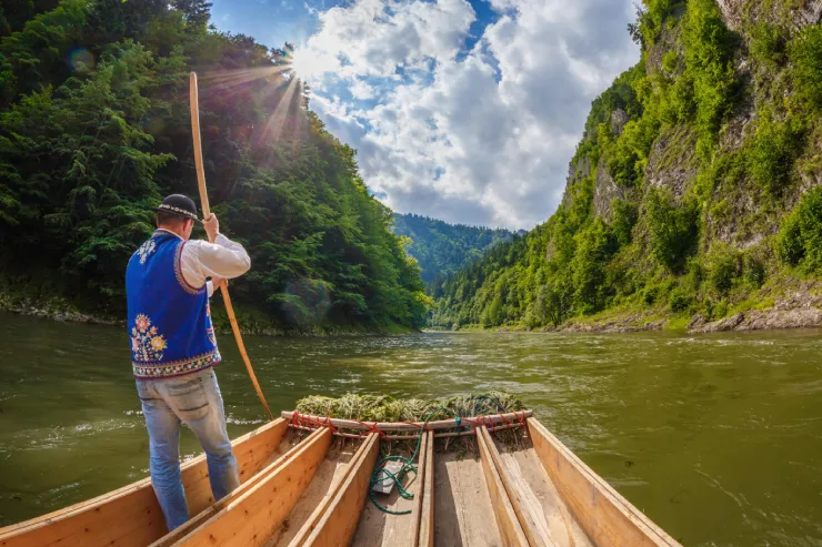 A wide-angle shot of a man wearing blue jeans, a traditional embroidered caftan over a white sleeved shirt, and a black round at with a string of white shells. He is holding a punt to navigate the raft in the foreground along a greenish river flowing fast into a gorge between rocks overgrown with greenery. Another slope in the background runs under copious clouds lit up by the sun.