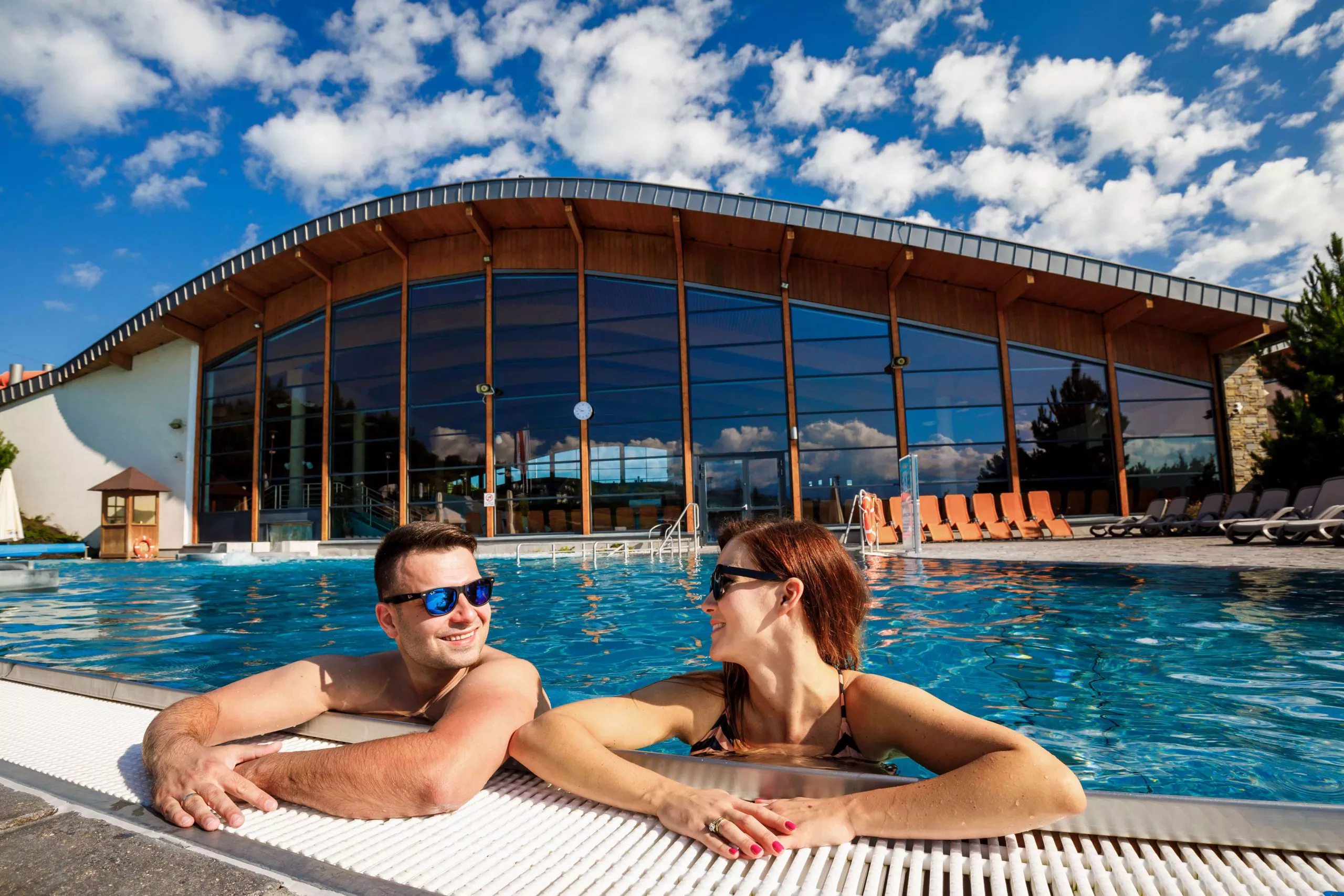 In the very foreground a happy couple in dark glasses are smiling at each other. Their arms rest on the edge of the pool, in which they are submerged up to their armpits. The Termy Bania building under blue sky with tufty white clouds provide the background.