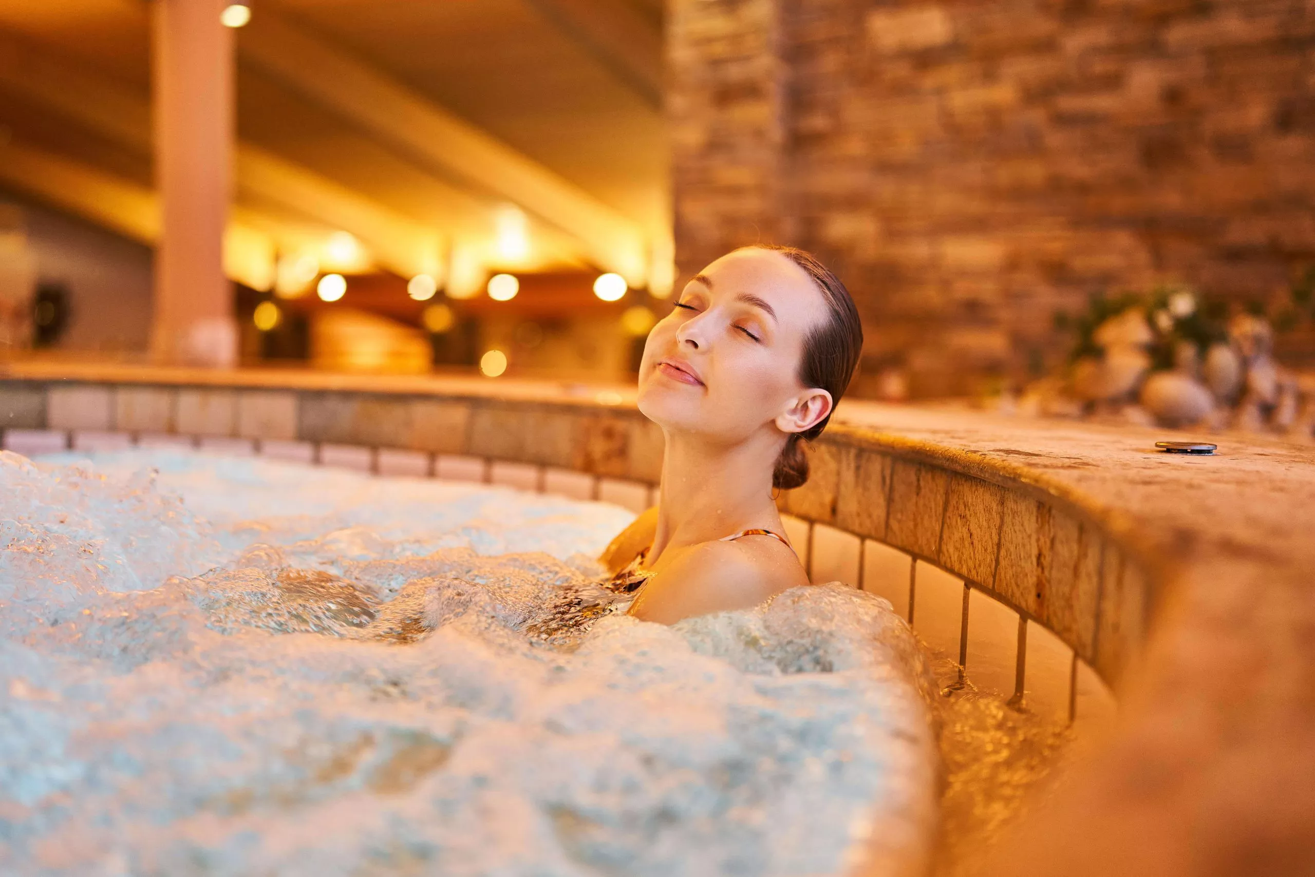 A relaxed young woman is resting in a bubbling jacuzzi-like pool, her eyes half-closed. The blurred background shows a stone wall and distant high ceiling supported by a column, lit with multiple lamps.