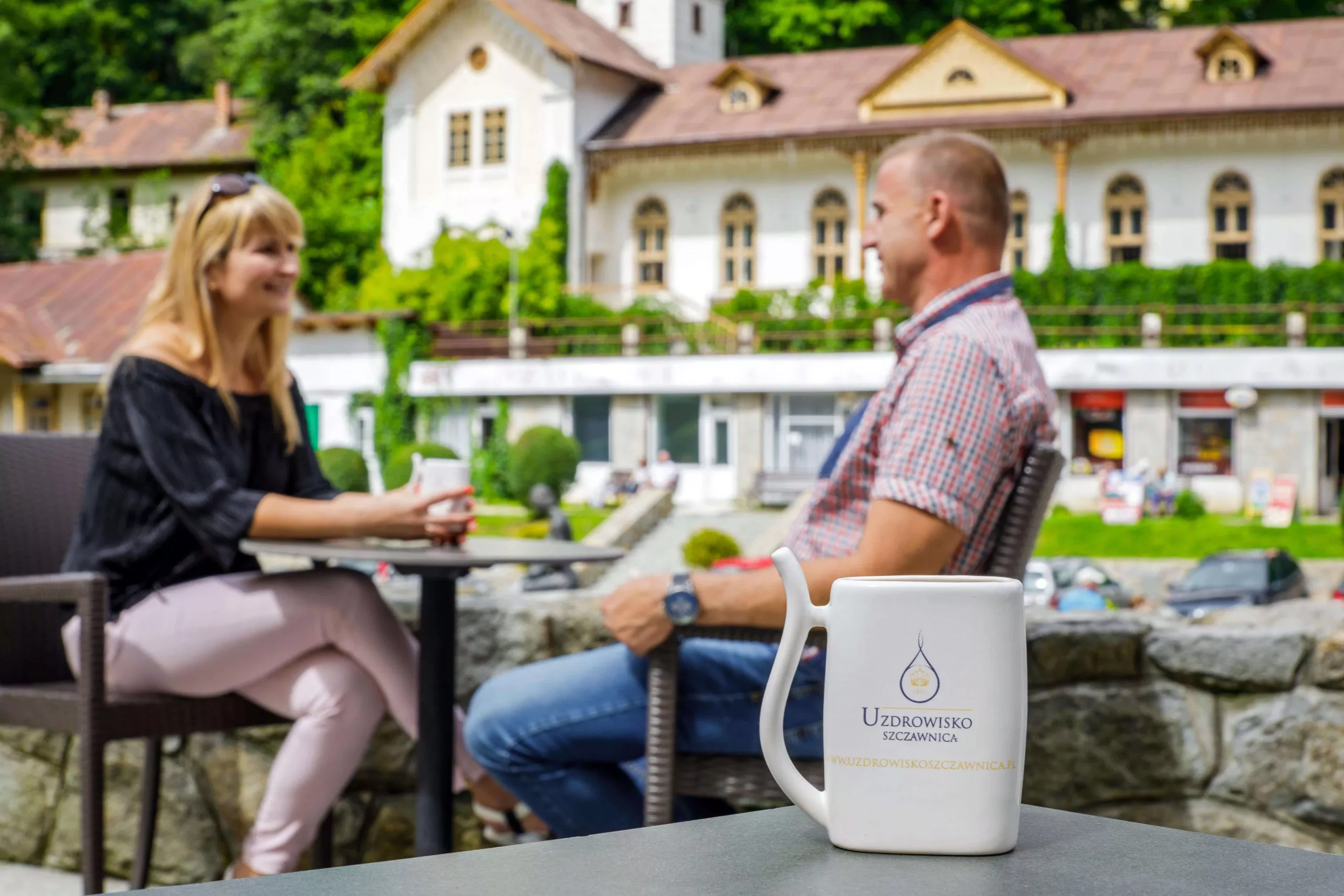 A white spa-cup with the logo and name of Szczawnica Spa provides the foreground for a middle-aged couple sitting at a black table. The blurred background consists of red-roofed white spa buildings among greenery.