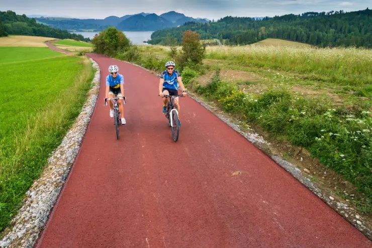 Blick auf zwei junge, behelmte Radfahrer, einen Mann und eine Frau, die auf einem rot gepflasterten Radweg fahren. In der Ferne kann man einen Teil des Czorsztyńskie-Sees und zahlreiche Hügel sehen. Rund um den Radweg wachsen verschiedene Sträucher und hohes Gras. Darüber erstreckt sich der blaue Himmel.