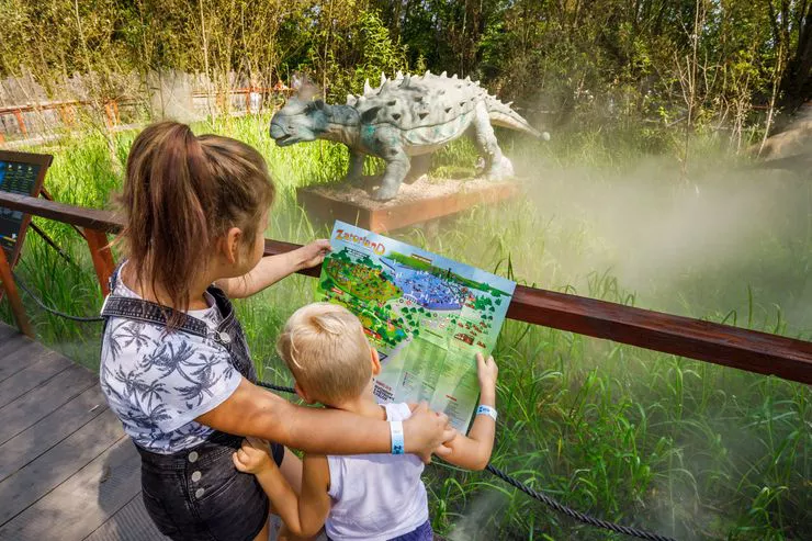 Blick auf zwei Kinder, die an der Absperrung stehen, die sie von der Dinosaurierfigur trennt. Die Kinder stehen rückwärts und lesen einen Flyer.