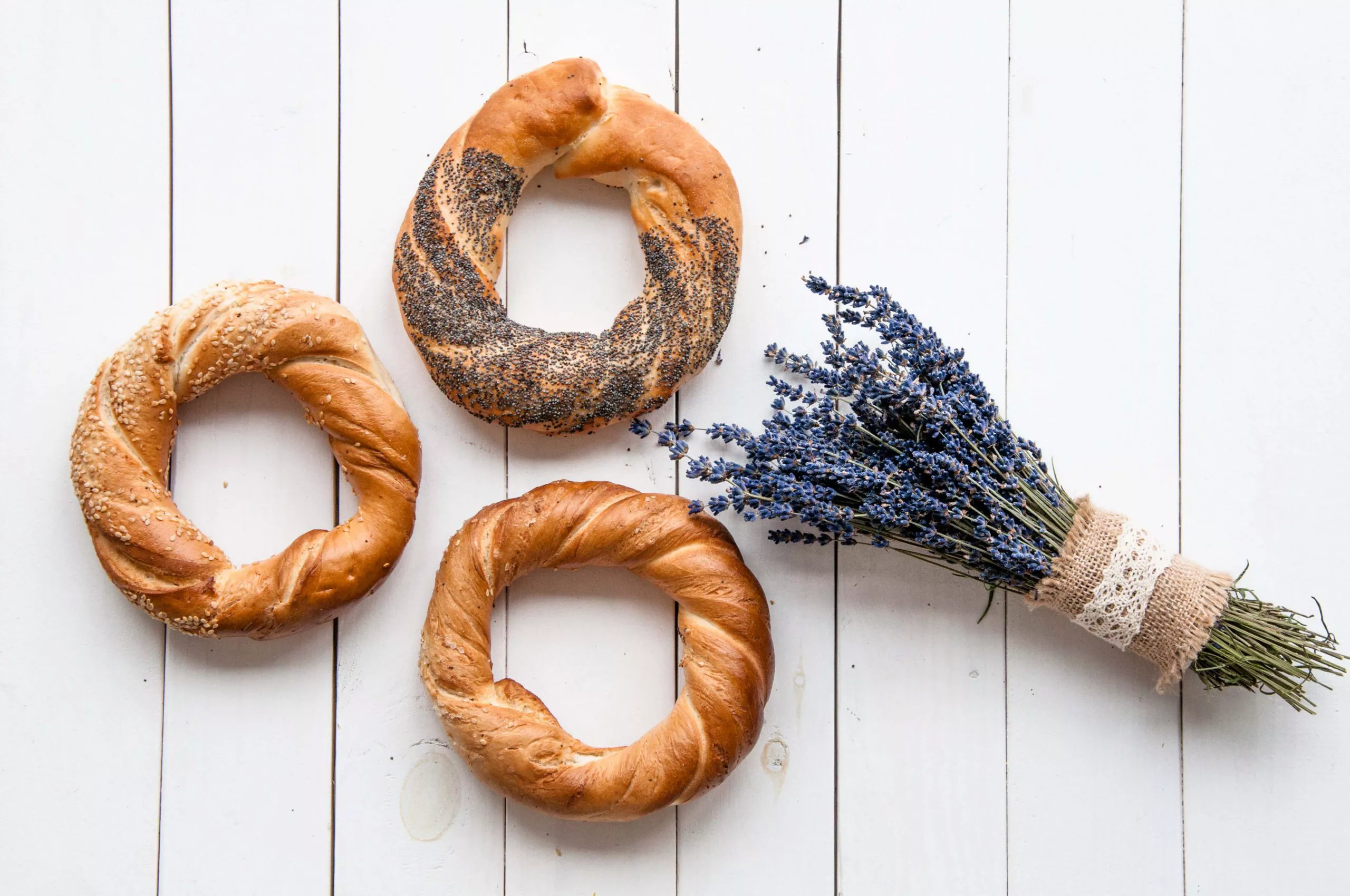 Three obwarzanki bread rings, the left one sprinkled with salt, the top one – with poppyseed, and the bottom one – with sesame, are arranged on white boards, with a rustic bouquet of tiny blue flowers on the right.