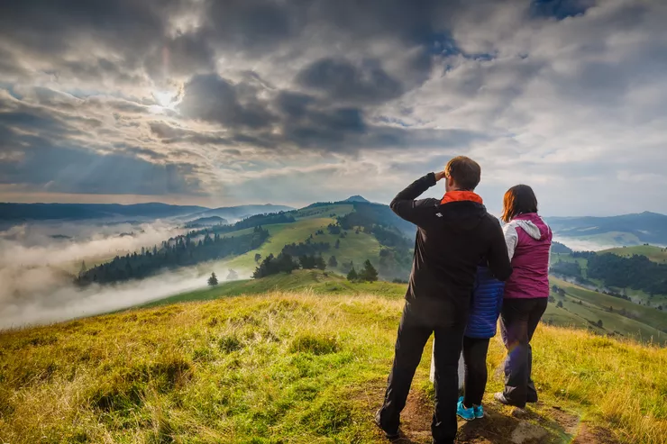 In the foreground, two adults with a child between them are standing on a broad grassy ridge admiring the mists crawling from the valleys below onto the farther sections of the ridge. The sky is overcast with heavyish clouds, yet some sunrays manage to pierce them.