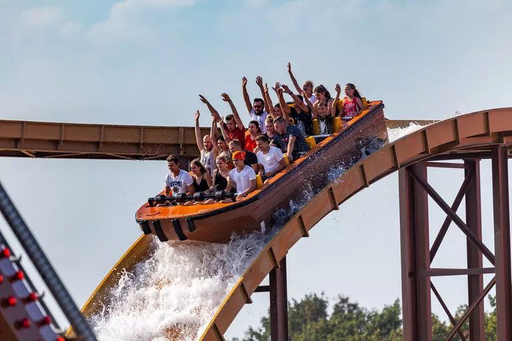 A large raised wooden water track with a boat going down a steep arch under an overcast sky. The people in the first row in the boat hold tight, while those sitting in the four rows behind them are holding their hands up in the air.