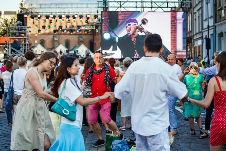 A group of people men and women dance in the street, holding hands. A high brightly lit stage stands in front of the brick building in the background. The TV wall on the right shows a man playing the trumpet.
