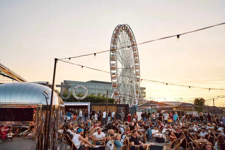 A thick young crowd are leisurely seated in deckchairs in the open at the foot of Forum Przestrzenie. In the background, a great Ferris wheel stands against a sunset.