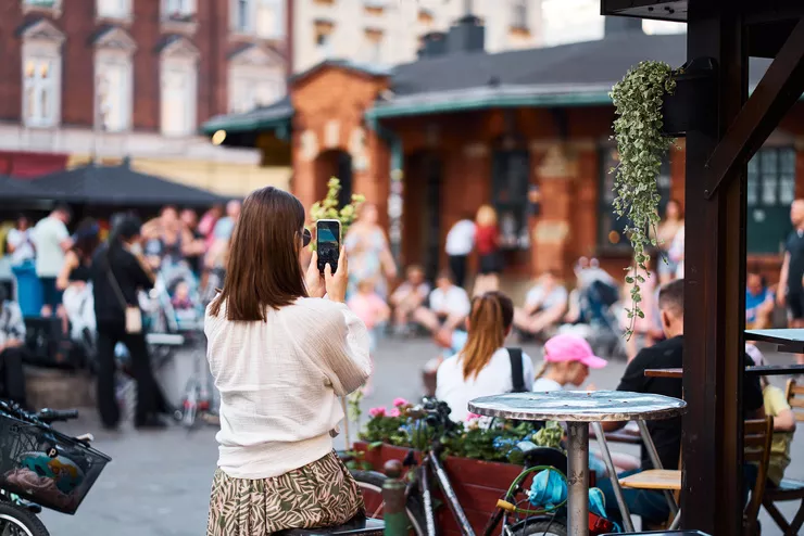 A young woman in a white blouse in the centre has raised her smartphone to take a shot of a low brick building in Nowy Square. There are blurred figures of people, some sitting at tables, some on the ground to her left, while the ones on the right stand in front of some stalls.