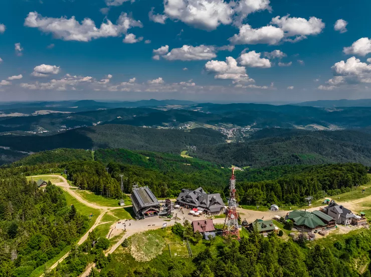 A bird’s eye view of the top of Jaworzyna Krynicka mountain with the upper terminal of the cable car topped with a steel truss viewing platform. Leading from it are tourist foot and bike paths. Standing to its left is a cluster of buildings catering for tourists. The whole stands on the first plane among forests that stretch on to the further ranges visible far away under cloudy skies.