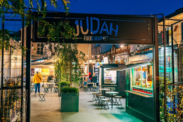 A view of the black gate bearing the inscription Judah Food Market. Standing within the perimeter of the fencing are food trucks with various delicacies standing around clusters of round tables with stools in the centre. Buildings can be seen behind and to the right of the square.