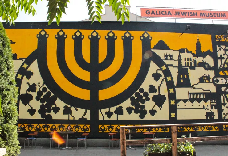 A mural on the wall of the Galicia Jewish Museum shows a Jewish candelabrum with seven branches against a background of various Kraków and Kazimierz buildings. Its dominant colours are dark yellow, black, and cream. There are a number of tables and chairs dwarfed by the size of painting standing along its bottom edge.