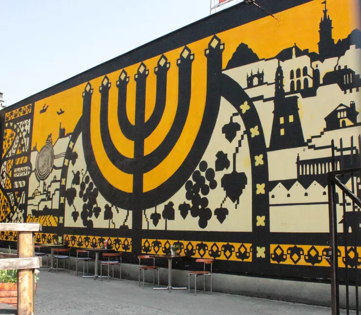 Blick auf ein Wandgemälde im Jüdischen Museum Galizien. Es stellt einen Kerzenständer und verschiedene Gebäude dar. Das Wandgemälde ist hauptsächlich in Gelb und Schwarz gehalten. Unter dem Wandgemälde befinden sich Tische mit Stühlen.