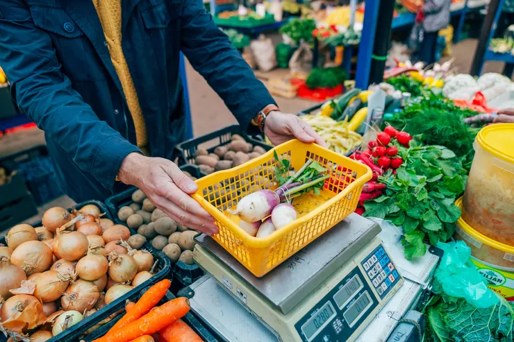 Vegetables are being sold in a market square from a stall with onions, potatoes, carrots, radishes, bell peppers, and cabbage. A man in a blue jacket on the left is holding a tray with four extra large radishes on his electric scales.