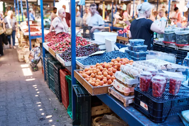 Lines of stalls with fruits, eggs, and vegetables running upwards from the bottom right corner. Arranged in flat boxes on them are peaches, blueberries, strawberries, and sweet and sour cherries. There are shoppers in the blurred background behind them.