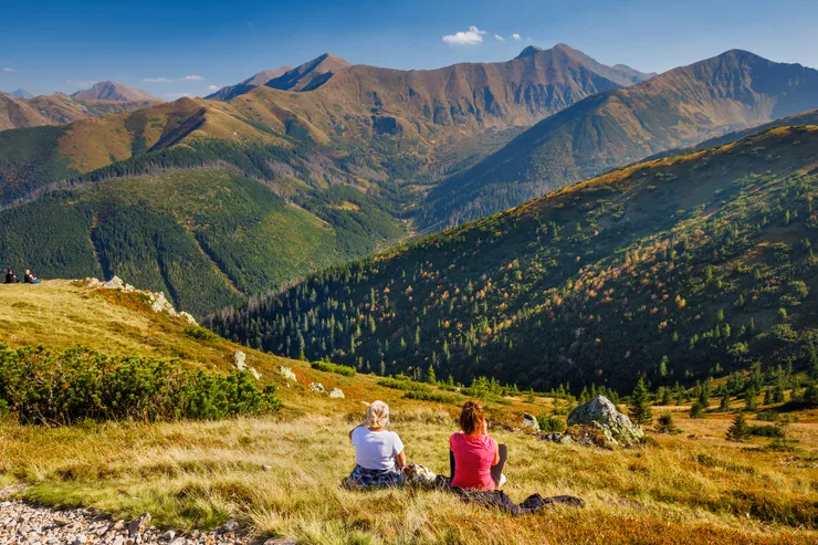 Blick auf zwei Frauen, die im Gras sitzen und die Gipfel der Westlichen Tatra bewundern. Im umliegenden Gras sind kleinere Felsensteine zu sehen. Es ist ein schöner sonniger Tag, der Himmel ist blau.