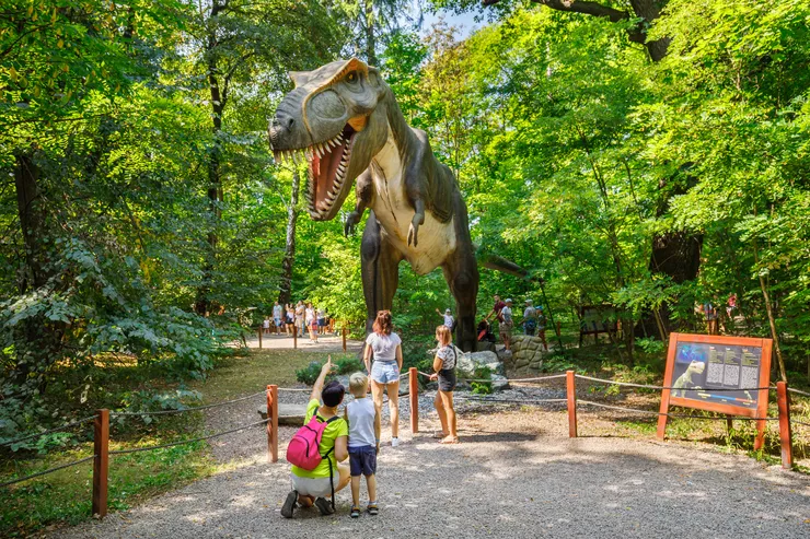 Two children standing by a barrier look dwarfed by a huge mechatronic tyrannosaurus in the centre of the picture behind it. Standing closer to us is a little boy, whose mother, wearing a bright green blouse and with a pink backpack, is pointing at the dinosaur with her finger. There are trees on both sides of the figure, and an information board on its far right by the barrier.