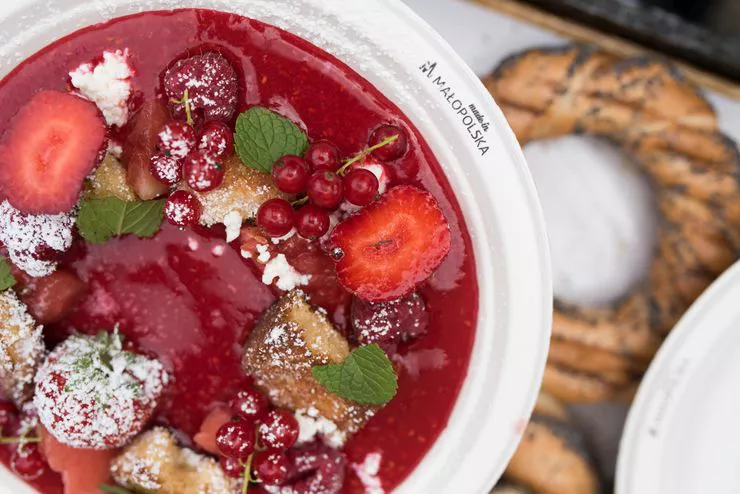 A plate of thick red fruit soup with chunks of strawberries, whole red currants, and mint leaves, slightly sprinkled with castor sugar. An out-of-focus Obwarzanek sprinkled with poppy seed lies below the plate’s rim, just below the inscription “Małopolska”.